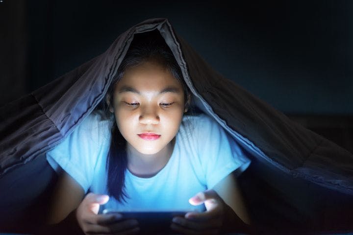 A young woman staring at her smartphone under her blanket in the dark.