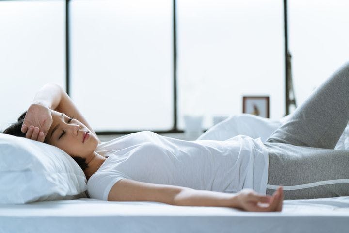Woman lying on a bed with her eyes closed while placing her left hand on her forehead