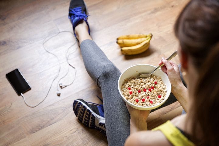 An over-the-shoulder shot of a woman holding a bowl of cereal while sitting on the floor next to a bunch of bananas.