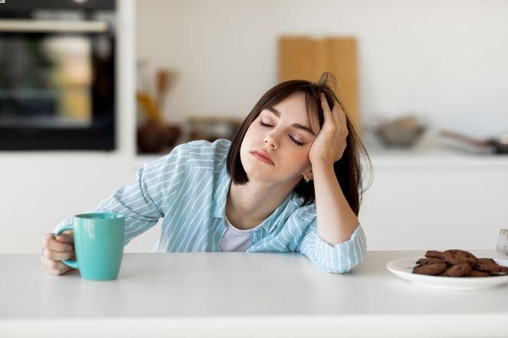 Woman falling asleep while holding a cup of coffee with a plate of chocolate cookies at the side