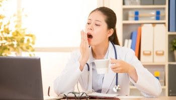A female doctor sitting on her work desk while yawning and holding a cup of coffee in one hand.