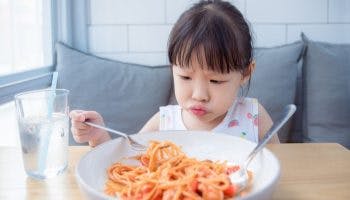 A girl with a frown on her face picking up spaghetti noodles from a bowl with a fork