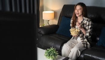 A woman sitting on a sofa while holding a bowl of popcorn while laughing at something she sees on TV. 