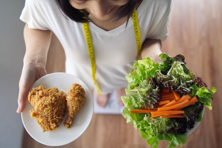 A top view of a woman standing on a weight scale while holding plates of fried chickens and salad.