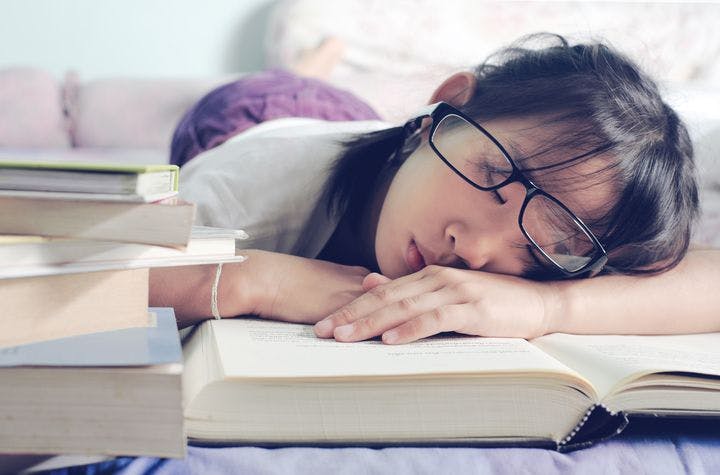 Woman with glasses asleep with her face and hands resting on a book