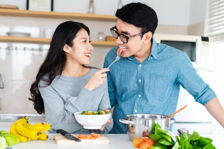 A woman feeding a man healthy food; fruits and vegetables spread on the kitchen island in front of them.