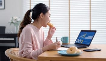 Woman eating a doughnut as she sits a table looking at her laptop