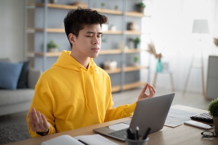 A young man sitting at his desk in front of his laptop while meditating with his eyes closed