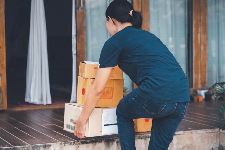 A woman is bending over to lift some heavy boxes outside her home