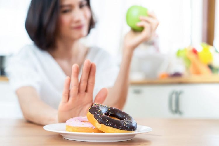 Woman holding an apple in her left hand as she lifts her right palm up to decline a plate of doughnuts on a wooden table