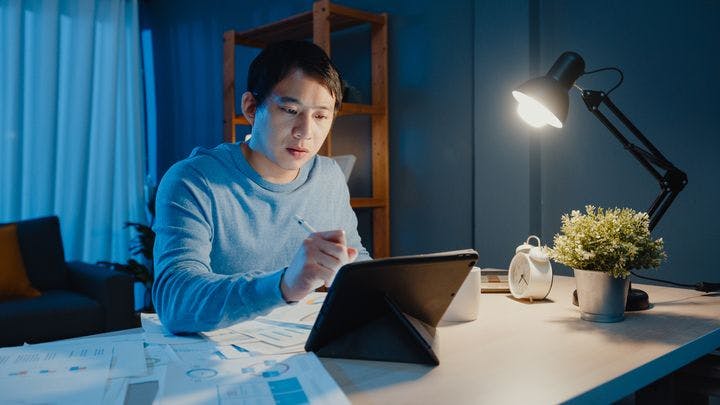 Man working at home at night in front of a tablet with papers on a desk.