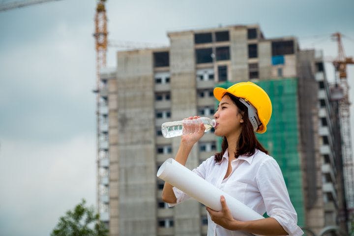 A woman wearing a protective hat is drinking water on a construction field