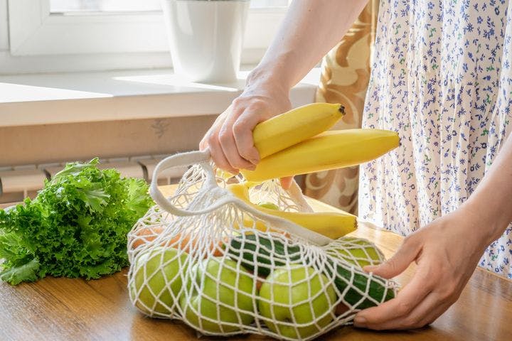 A partial view of a woman's hands pulling fruits and vegetables out of a knitted bag.