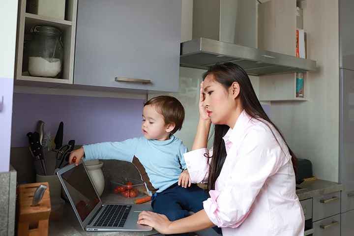 Woman working from home on laptop in the kitchen while also tending to her toddler.