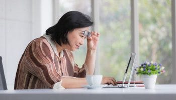 Woman lifting her eyeglasses to look at her laptop while sitting at a table