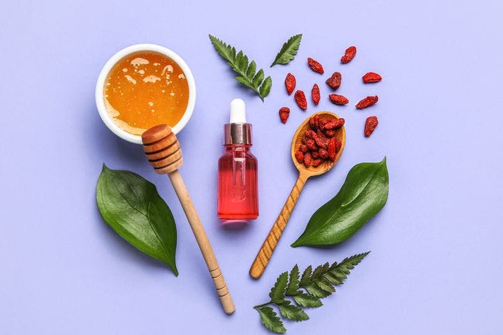 An aerial view of a bottle of skincare product surrounded by a spoon of goji berries, a bowl of honey and leaves against a blue background.