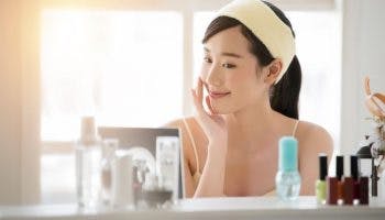 Asian woman touching one cheek while smiling and looking at skincare products on the table in front of her