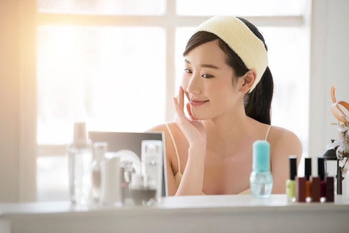 Asian woman touching one cheek while smiling and looking at skincare products on the table in front of her