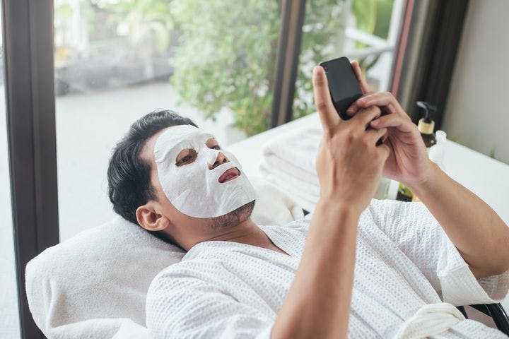 A man wearing sheet mask and playing on his phone while laying down on a bed