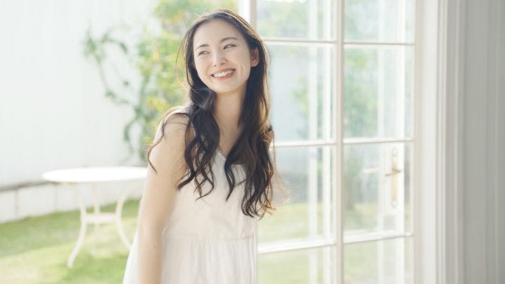 A woman smiling while standing beside a tall glass window with the view of a garden behind her.