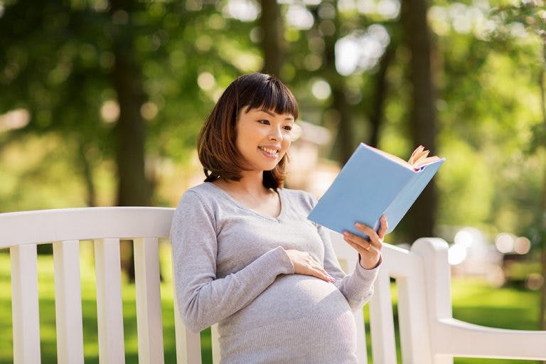 A pregnant woman sits in a park reading a book