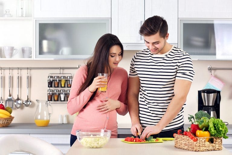 A pregnant woman looking at her husband chopping vegetables in the kitchen