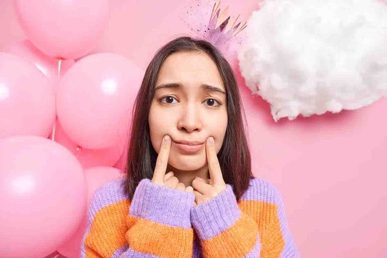An Asian woman stands in front of a wall decorated with balloons and projects a fake smile by pressing her two fingers to her mouth