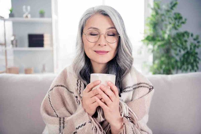 An older Asian woman smiling while drinking from a mug on a couch