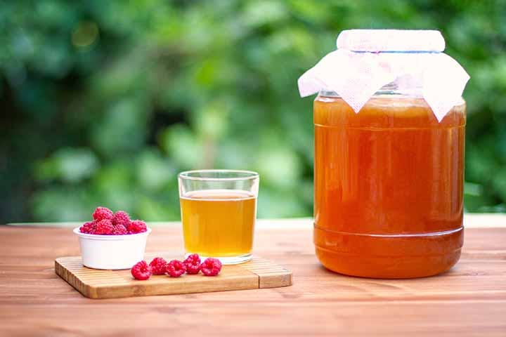A large kombucha in a glass jar, a small glass containing kombucha and a bowl of raspberries on a wooden table