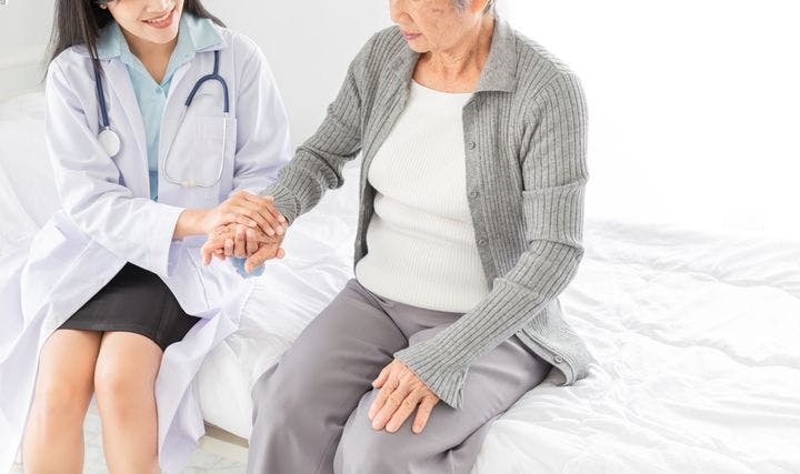 A doctor holding an elderly woman’s hand as they sit up on the edge of a bed