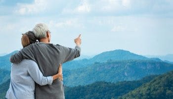 Man and woman embracing as the man points with his right finger as they look at a view of mountaintops
