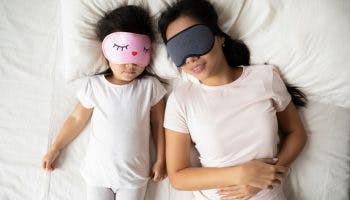 An overhead shot of a mother and her daughter sleeping side by side while wearing eye masks.