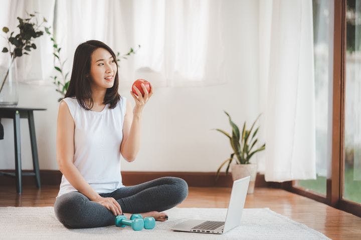 A woman sitting cross-legged on the floor while holding an apple; small hand weights and a laptop lying around her.