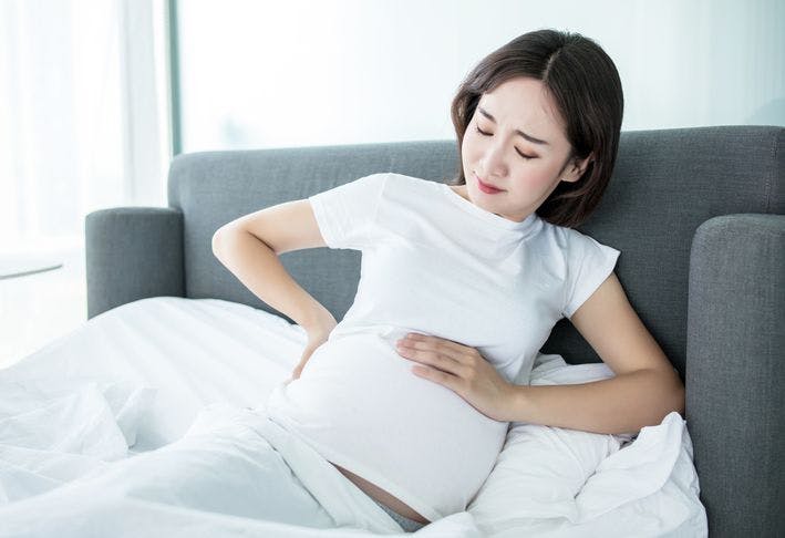 A pregnant woman sitting up in bed while holding her abdomen and lower back.