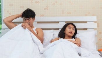 Man with a confused facial expression as he looks under the covers and a displeased woman looking to her side as they both lie in bed