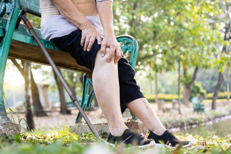 An elderly woman sitting on a bench holding her knee and experiencing arthritis pain