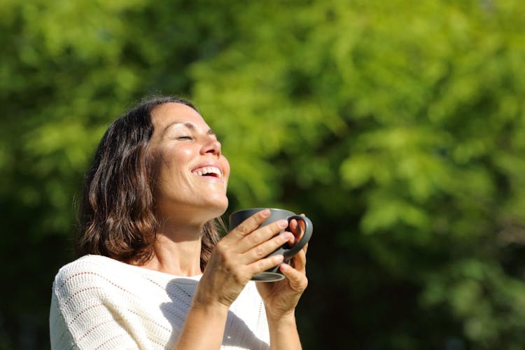A woman smiling outdoor near trees holding a cup