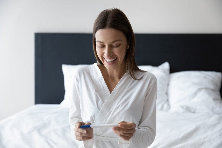 A woman reacts happily to the result of a home pregnancy test kit while sitting in bed