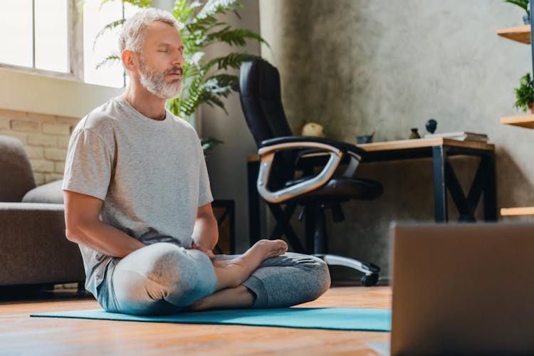 A middle age man meditating on a blue yoga mat in front of a laptop