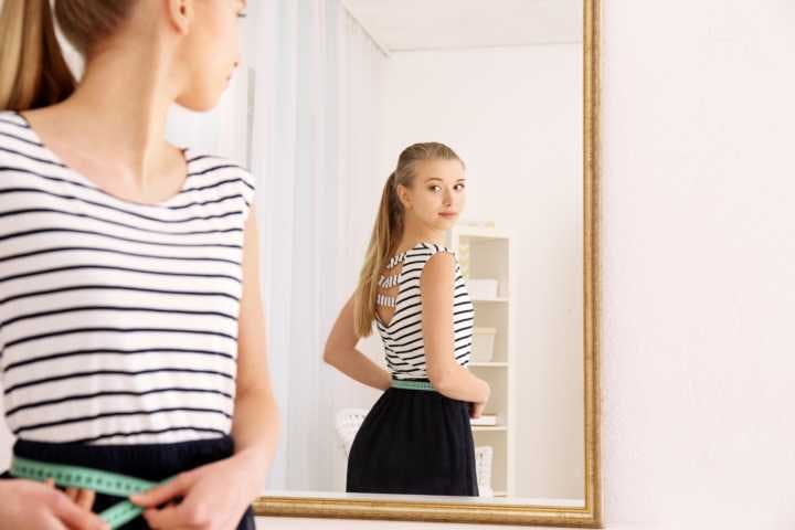A beautiful skinny woman looking at herself in the mirror while measuring her waist
