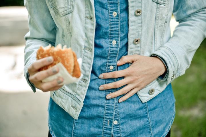 A woman touching her stomach in pain as she eats a hamburger on a street