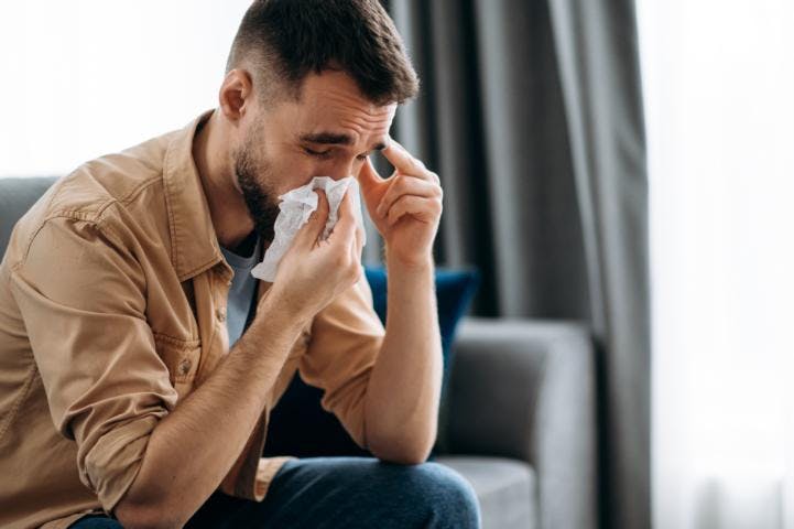 A young caucasian man is having sinusitis headache while blowing his nose with a tissue