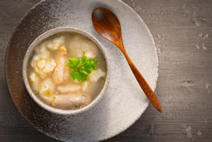 A bowl of wonton soup with parsley on a wooden table top with a wooden spoon next to it