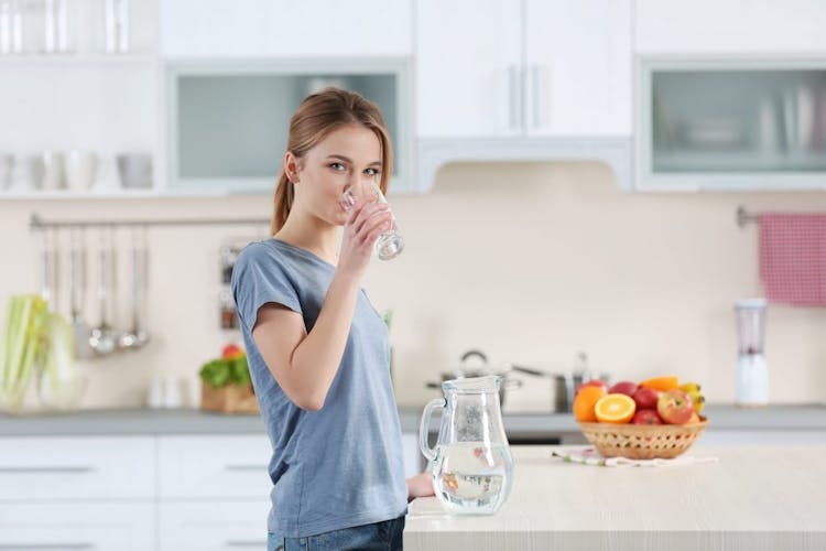 A young woman standing in her kitchen drinking water 