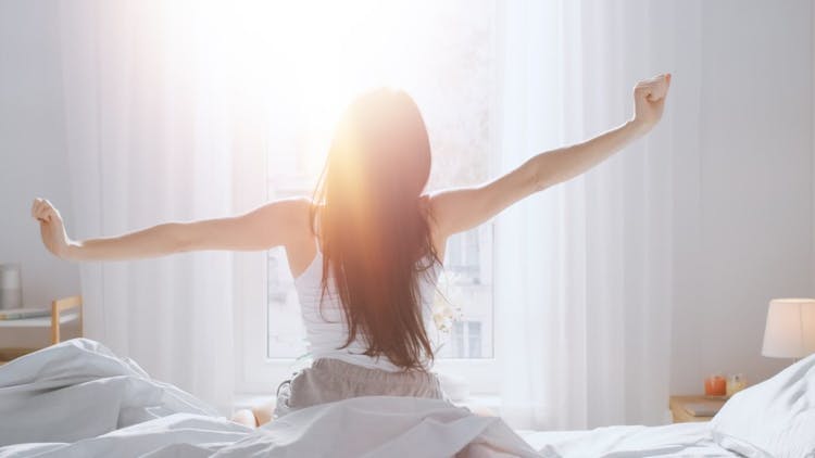 A photo of a young woman waking up and stretching as the morning sun comes through her window after a good night's sleep