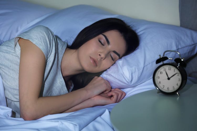 A young woman sleeping next to an alarm clock that reads approximately 11 pm