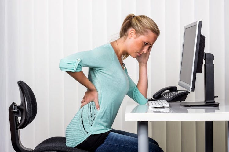 A blonde woman sitting at a computer desk holding her lower back in pain