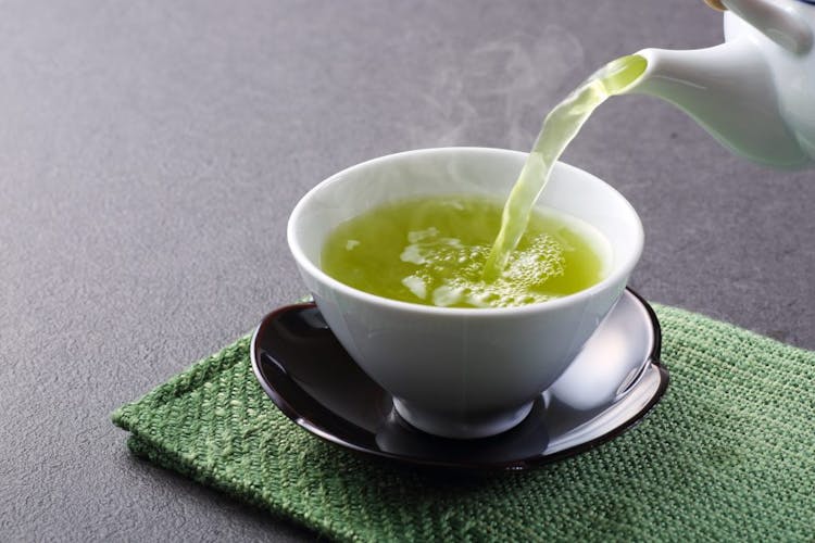 Hot green tea being poured from a tea kettle into a white tea cup