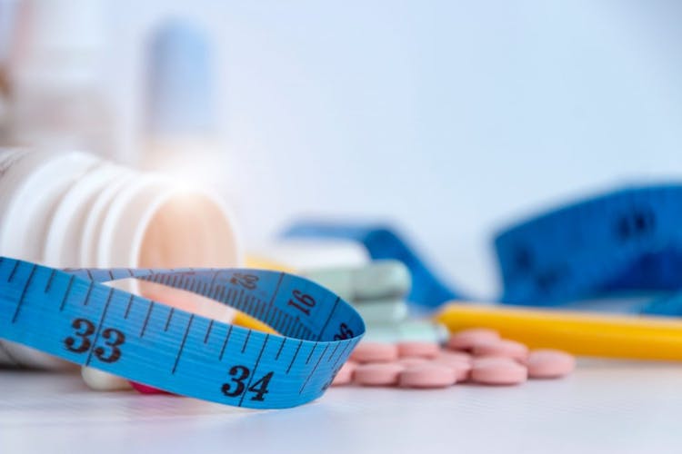 An image of pink weight loss pills from a bottle and blue tape measure to measure waist circumference 