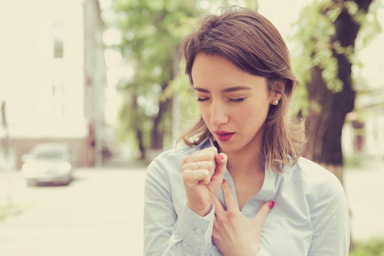 A woman coughing while putting her hand on her chest outdoor.